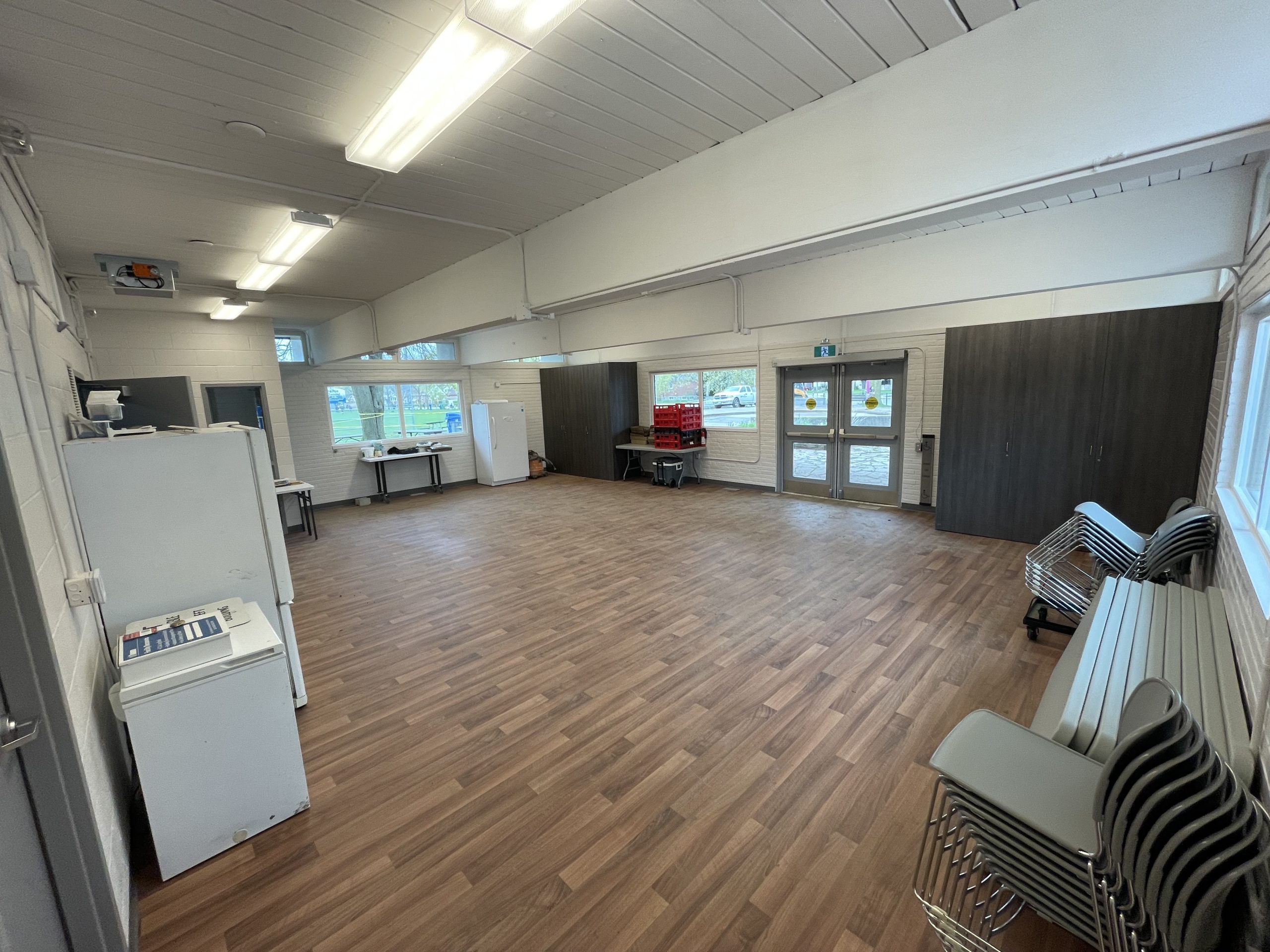 A large community room inside the clubhouse showing an open space with windows and stacked chairs and tables along the wall.