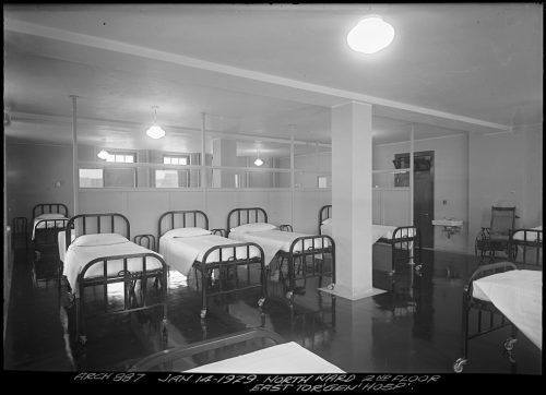 Image depicts monochrome photograph of interior of hospital ward