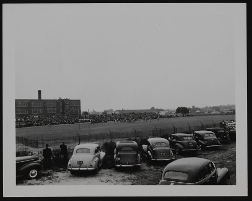 Image depicts monochrome photograph of automobiles parked on perimeter of football field with crowd watching game