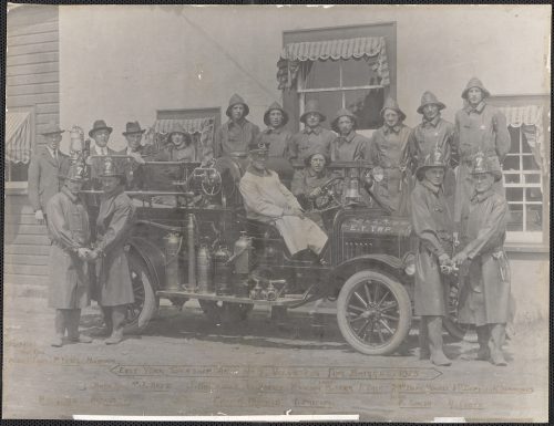 Image depicts monochrome photograph of fire engine surrounded by volunteer fire fighters