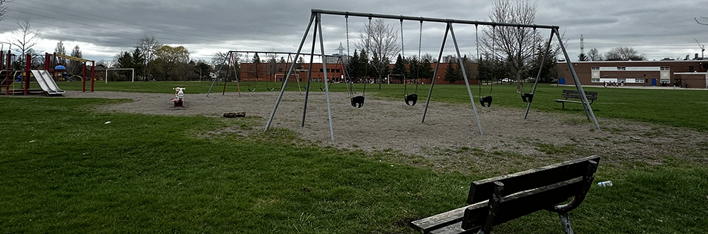 Huntsmill Park Playground with a bench facing the swing set in the foreground, and another swing set, spring toy and slides in the background.