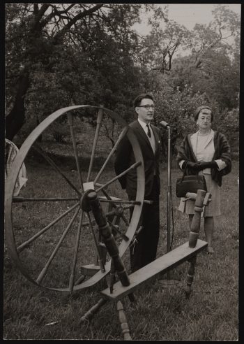 Image depicts monochrome photograph of two people standing beside an oversized spinning wheel