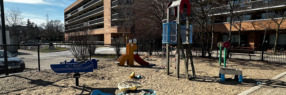 A playground with various stand-alone play equipment on top of sand. An apartment building is in the background.