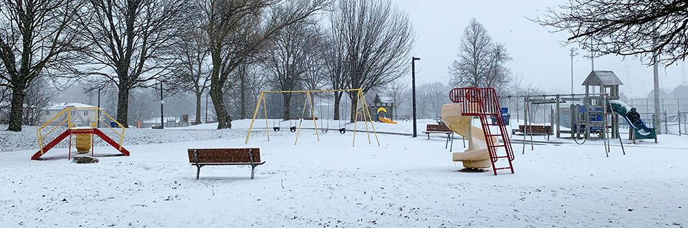 McNicoll Park Playground in the winter with snow. It shows a bench, small structure and spiral slide in the foreground, and a swing set and senior play structure in the background.
