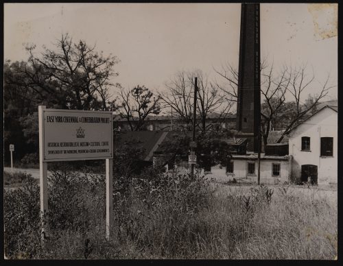 Image depicts monochrome photograph with chimney and Victorian two storey industrial buildings in background , and sign in foreground