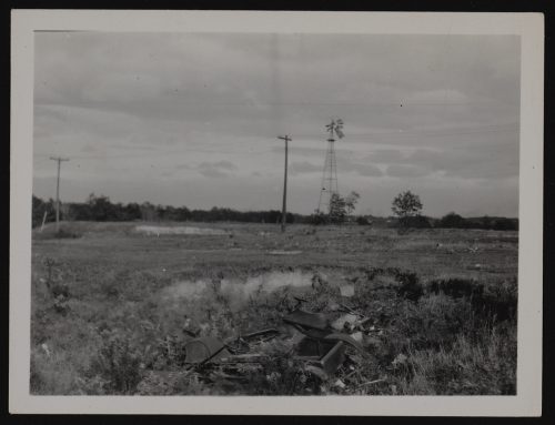 Image depicts monochrome photograph of field, with windmill hydro poles in background, and dumped garbage in foreground