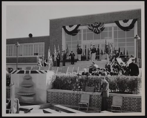 Image depicts monochrome photograph of group of people standing on front steps of municipal building