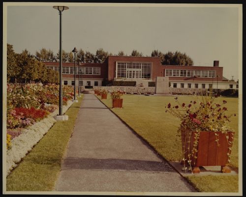 Image depicts colour photograph of municipal building with lawn and flower beds in foreground