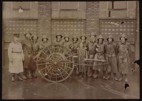 Image depicts monochrome photograph of group of fire fighters around hose wagon