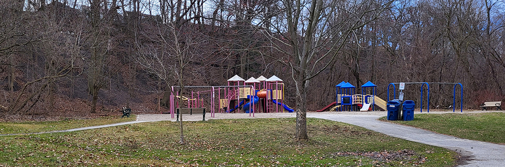 Smythe Park Playground in the fall showing a path leading to the playground area with a bench, a large play structure, a small play structure and a swing set. The playground is surrounded by mature trees.