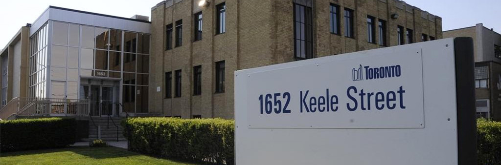 A brown brick building with floor to ceiling glass entryway with a sign on the green lawn that reads 1652 Keele Street, with a garden of small yellow flowers in front of it.