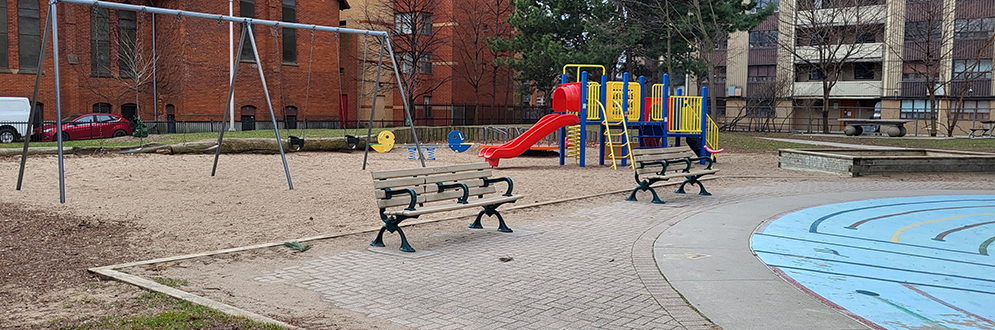 Masaryk Park Playground with two benches facing the splash pad in the foreground and a swing set, see-saw and play structure in the background.