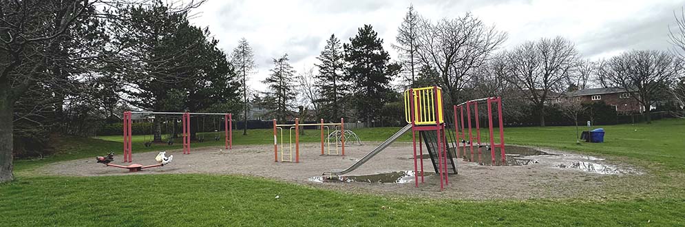 Highland Heights Park Playground showing play equipment in red and yellow on top of sand. The playground area is round in shape and surrounded by grass and trees.