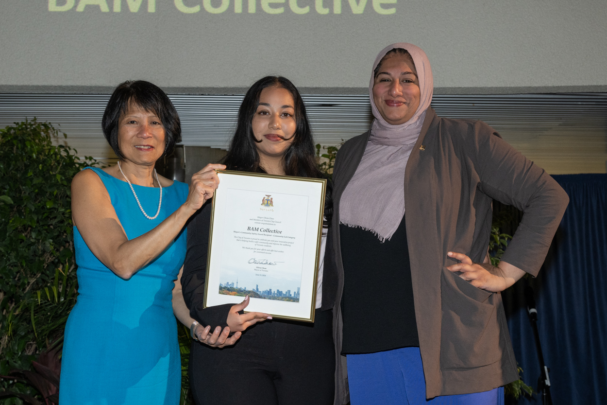 A women smiling for the camera with Mayor Olivia Chow and Deputy Mayor Ausma Malik holding up a framed award.