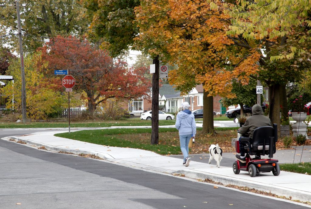 Two people, one of whom is using a power wheelchair, travel along a newly constructed sidewalk in a residential neighbourhood. 