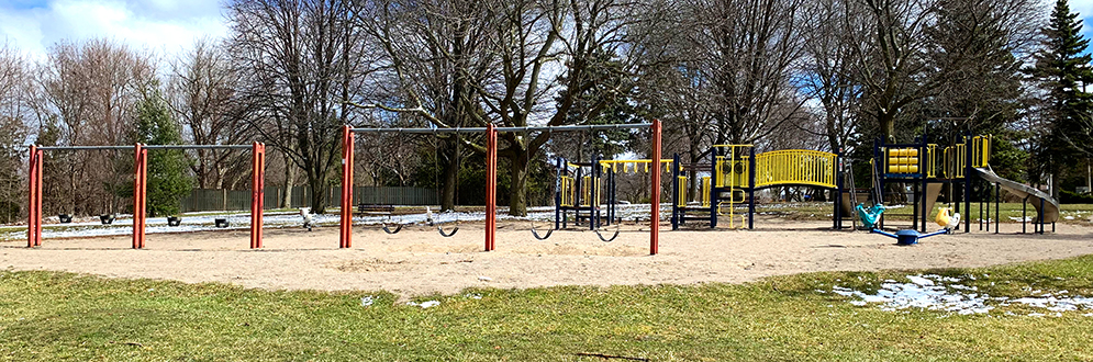 Woburn Park playground looking towards the east from the open lawn area to the west of the playground. The playground has mature trees along the east side, a walkway to the south, and open lawn to the north and west.