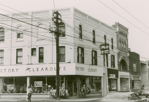 A black and white photo of the three-storey, mixed-use, rectangular building at the corner of Dundas Street West and Mavetry Street (photo taken facing west). The row of buildings along Dundas Street West recedes into the background to the right of the photo. 