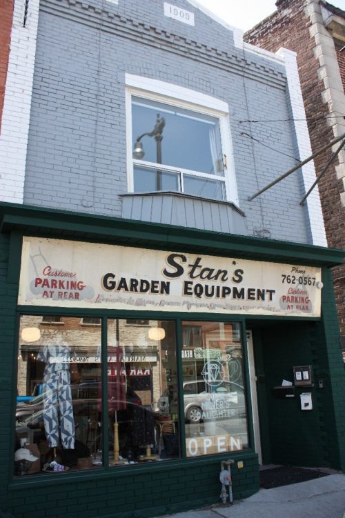 Photo shows a two-storey, mixed-use building with an angled storefront at street level and a painted sign reads: Stan’s Garden Equipment. The brick upper storey has a parapet with a date that reads 1909.