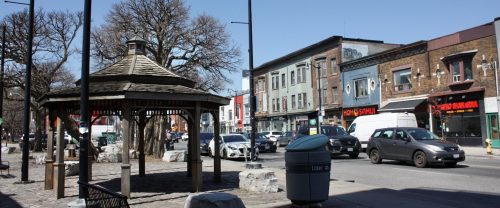 Facing northwest, a sunny view of the gazebo fronting onto Dundas Street West. Behind the gazebo are two large deciduous trees without leaves. Across the street from the gazebo is a row of two-storey to three-storey commercial and mixed-use buildings in multiple materials and colours. 