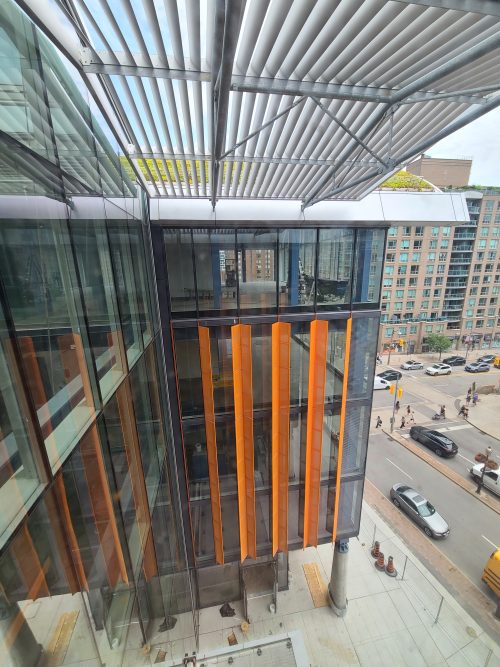 The side of a glass building with orange siding showing a street below with cars and pedestrians.