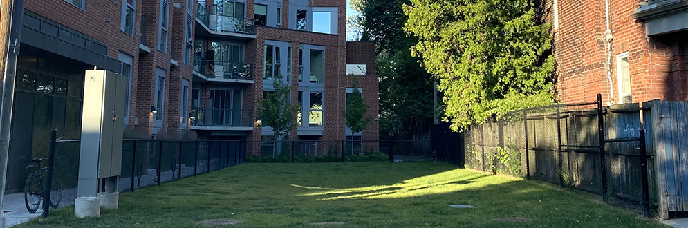 The park location from the sidewalk showing a rectangular patch of grass in-between a condo on the left and a house on the right.