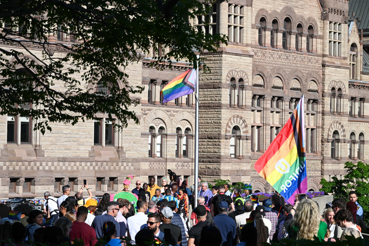 A large group of people gathers around a flag pole flying the Progress Pride Flag which features rainbow colours.