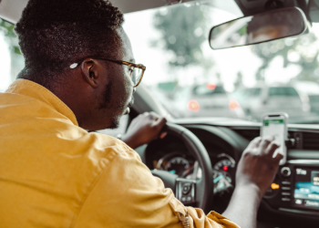 A man in a yellow shirt typing information into a mobile phone as he sits in the driver's seat of a car.