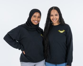 2 women in black sweatshirts standing in front of a white backdrop.