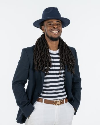 A Black man dressed in black and white standing in front of a white backdrop posing for the camera.