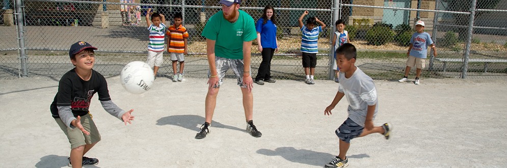 A child is trying to catch a ball, while another child is running to touch the base in a game of Baseball Soccer, while a staff is supervising.