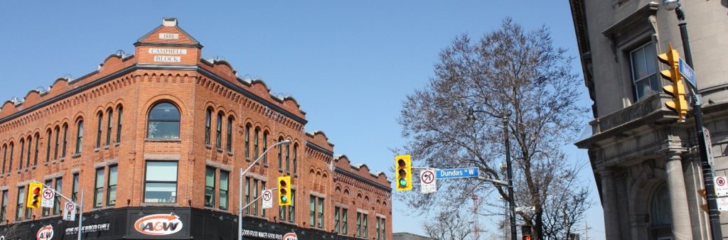 Facing northwest up Keele Street, the photo shows the intersection with two prominent corner buildings. To the left, the three-storey red brick Campbell Block building with chamfered corner, rows of round arch and flat arch windows, and decorative parapet. To the left is the rounded corner entrance of the former Bank of Toronto, a Neo-Classical building with ashlar veneer.