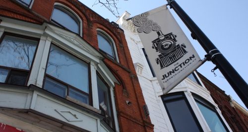 Photo shows a skyward view of the upper storeys of two mixed-use historic buildings fronting onto Dundas Street West taken from street level: the building to the left of the photo is red brick with a wood bay window painted pale blue surmounted by three arched windows. The adjacent building to the right is painted white with a bay window that rises two storeys up to a decorative cornice. A signpost is in the foreground with the Junction BIA banner: a black graphic image of a train on a white background.