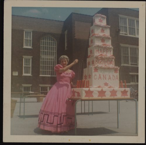 Image depicts colour photograph of East York Mayor True Davidson wearing a pink dress standing next to a giant birthday cake celebrating the centenary of confederation