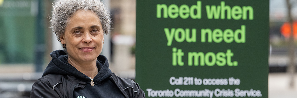 Close up of a person posing for the camera standing next to a sign that reads When You Need It Most, Call 211, Toronto Community Crisis Service