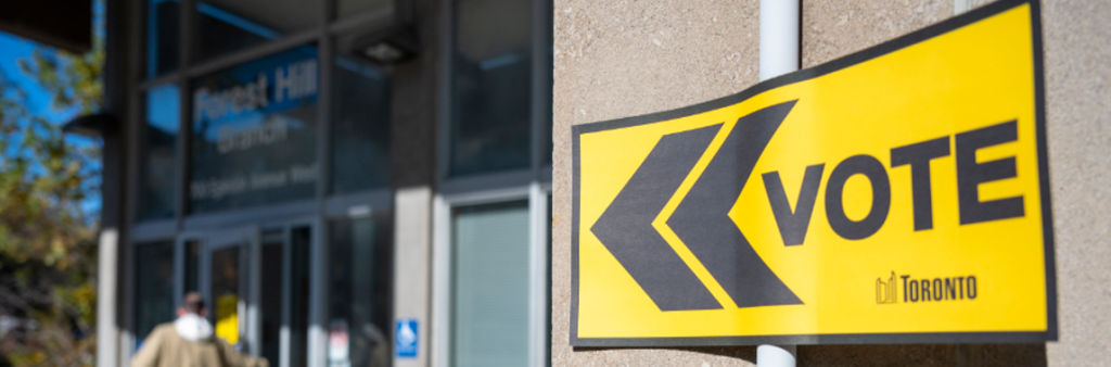 In the foreground of this image there is an official City of Toronto yellow and black vote directional sign posted out front of a voting place on a sunny day. A person walking into the voting place is blurred in the background.