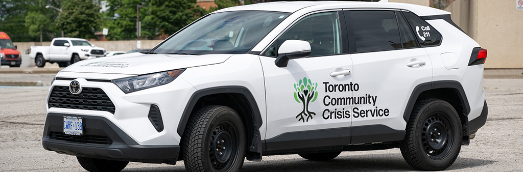 A white vehicle with a logo on the side reading Toronto Community Crisis Service as well as a logo in the rear window reading Call 211.