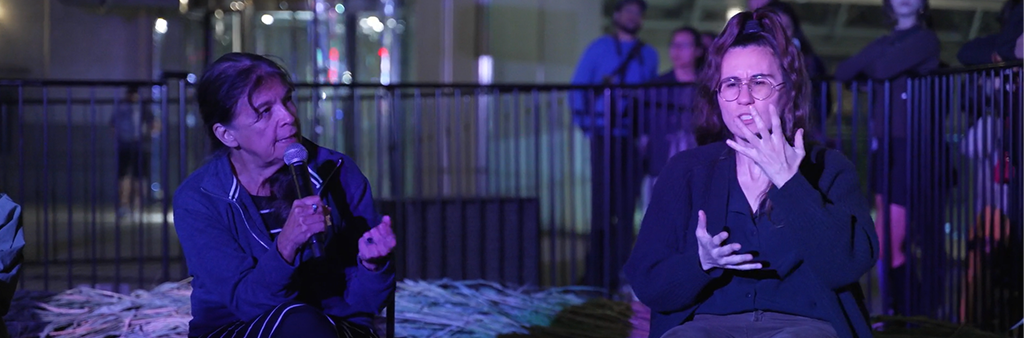 Two seated people at night talking. Person on left holding a microphone and person on right doing ASL interpretation with hands in front of face and chest.