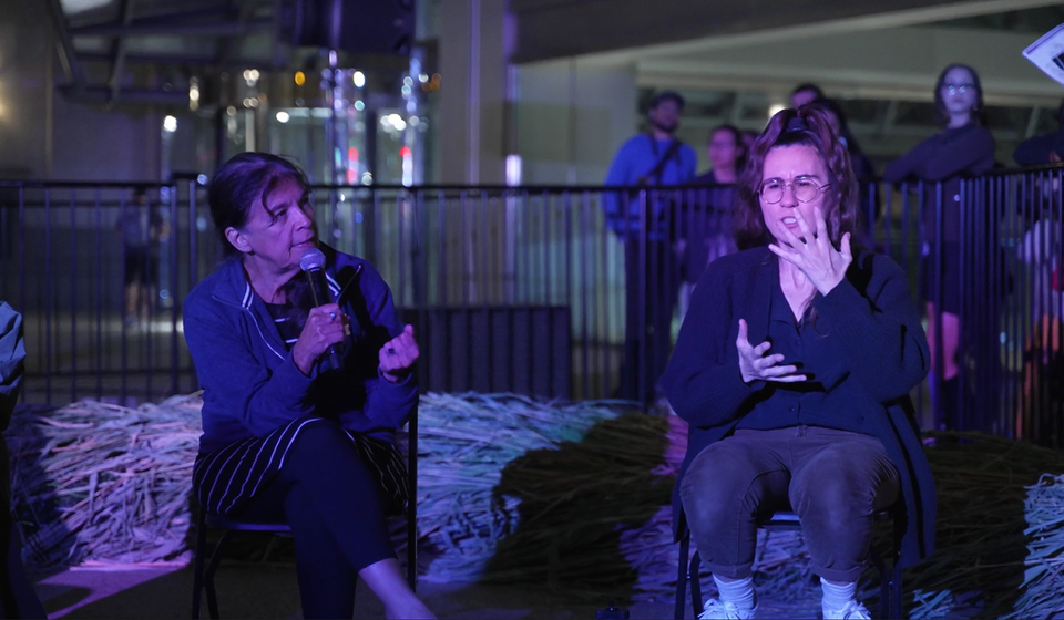 Two seated people at night talking. Person on left holding a microphone and person on right doing ASL interpretation with hands in front of face and chest.