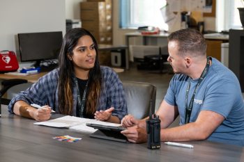 Two people seated at a table talking with a tablet, walkie-talkie and notebook on the table.