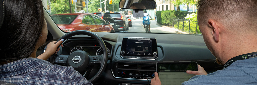 The backs of two people sitting in a vehicle, one holding a tablet with a map on it, looking out the windshield.