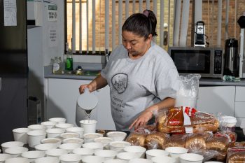 A woman in a kitchen pouring water from a kettle into many paper bowls.