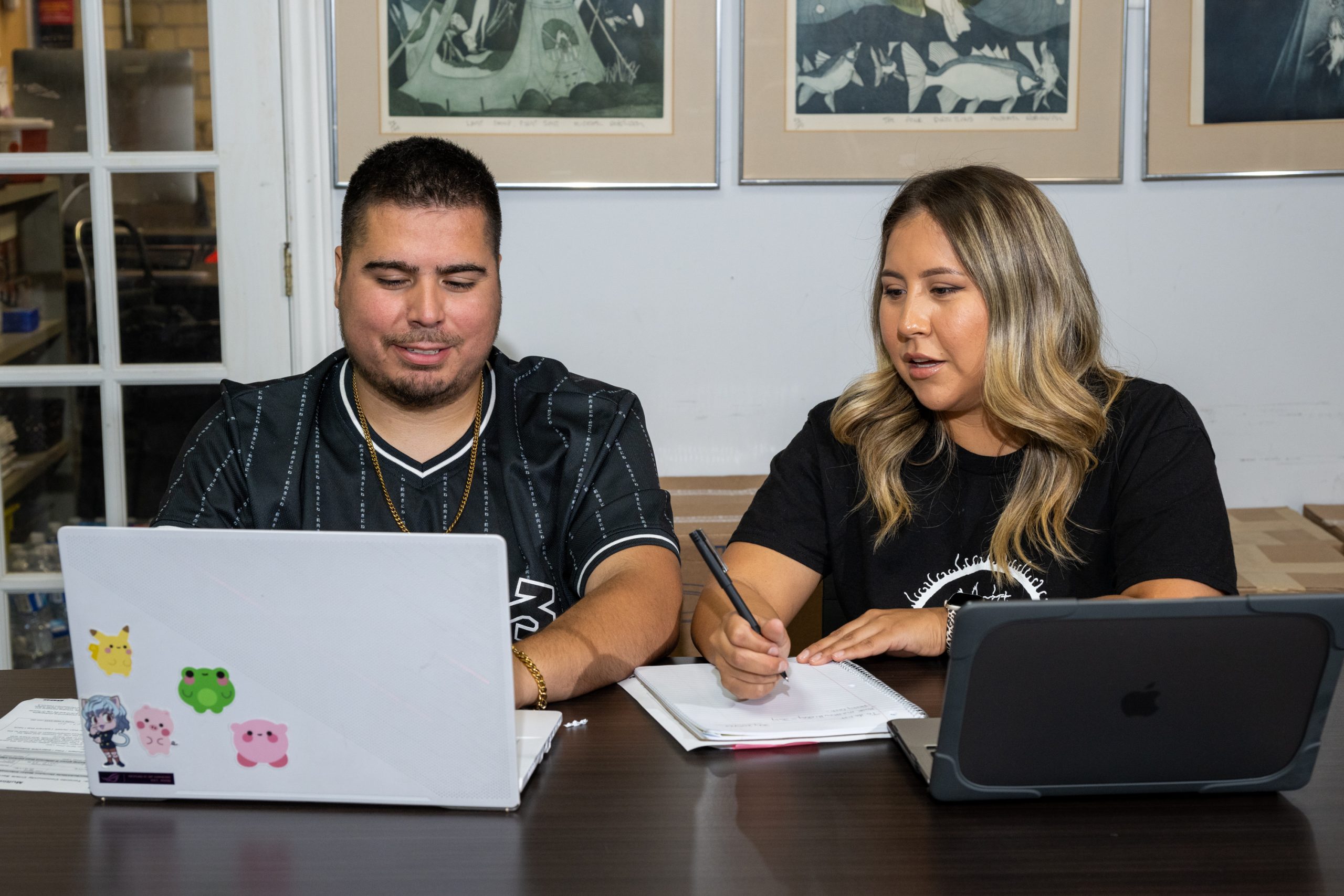 Two people working together at a table with laptops and taking notes.