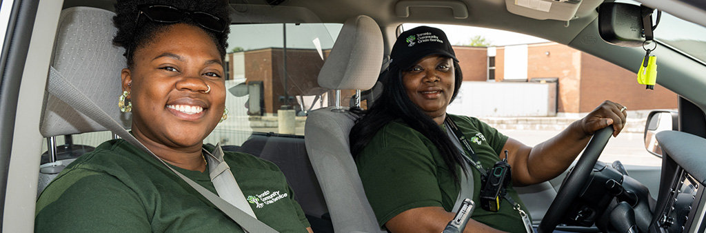 Two Black women sitting in a vehicle smiling for a photo wearing green Toronto Community Crisis Service shirts.