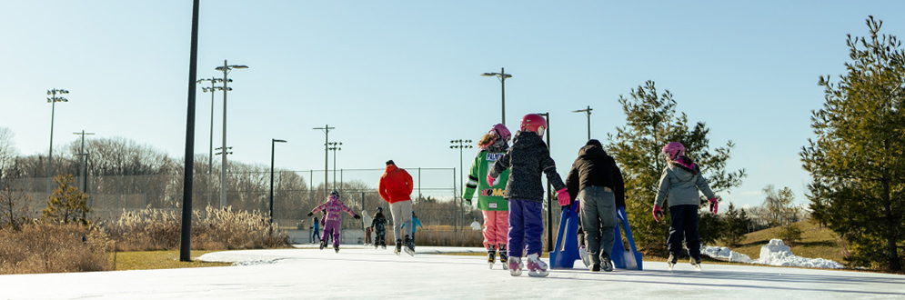 A group of people, including children, are ice skating on an outdoor skating trail on a clear, sunny day. The skaters are dressed in winter clothing and some children are using skating aids for balance. The skating trail is surrounded by trees and sports fields, and tall light poles line the trail