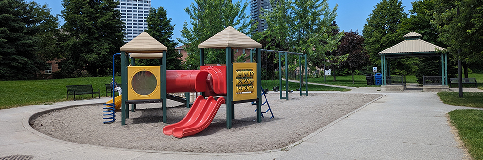 A small playground on sand with a play structure in red, yellow and green, and a swing set, circled by a concrete path with a pergola in the background.