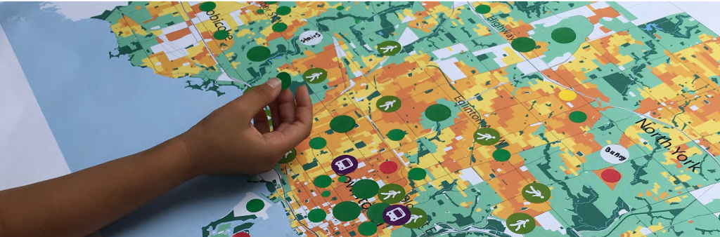 An overhead view of a person's hand placing a green marker on a detailed map of Toronto. The map highlights different areas in various colors, representing data on parks and land use. Several circular markers, representing different points of interest, are spread across the map, with labels for notable regions like North York and major streets such as Eglinton Avenue