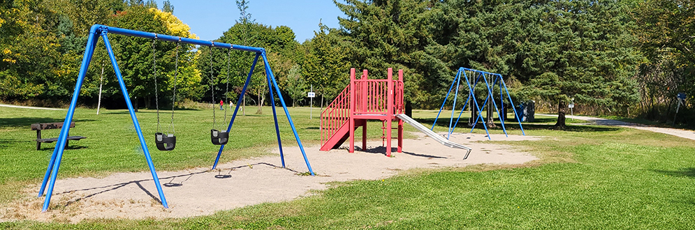 : A playground with a swing set with two toddler swings next to a small slide and another swing set with belt swings. The playground is on top of sand with grass and mature trees surrounding it.