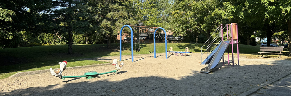 From left to right, the image shows a green teeter totter, swingset with one tot swing and one belt swing, a blue teeter totter, and a red slide. There is sand surfacing beneath all equipment.