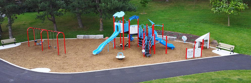 New playground area looking south, the play equipment is a mix of bright blue, red and light grey and has engineered wood fibre safety surfacing. At the front, the playground is surrounded by a new asphalt walkway. At the back is armourstone seating with a new garden and evergreen trees on a grassy slope.