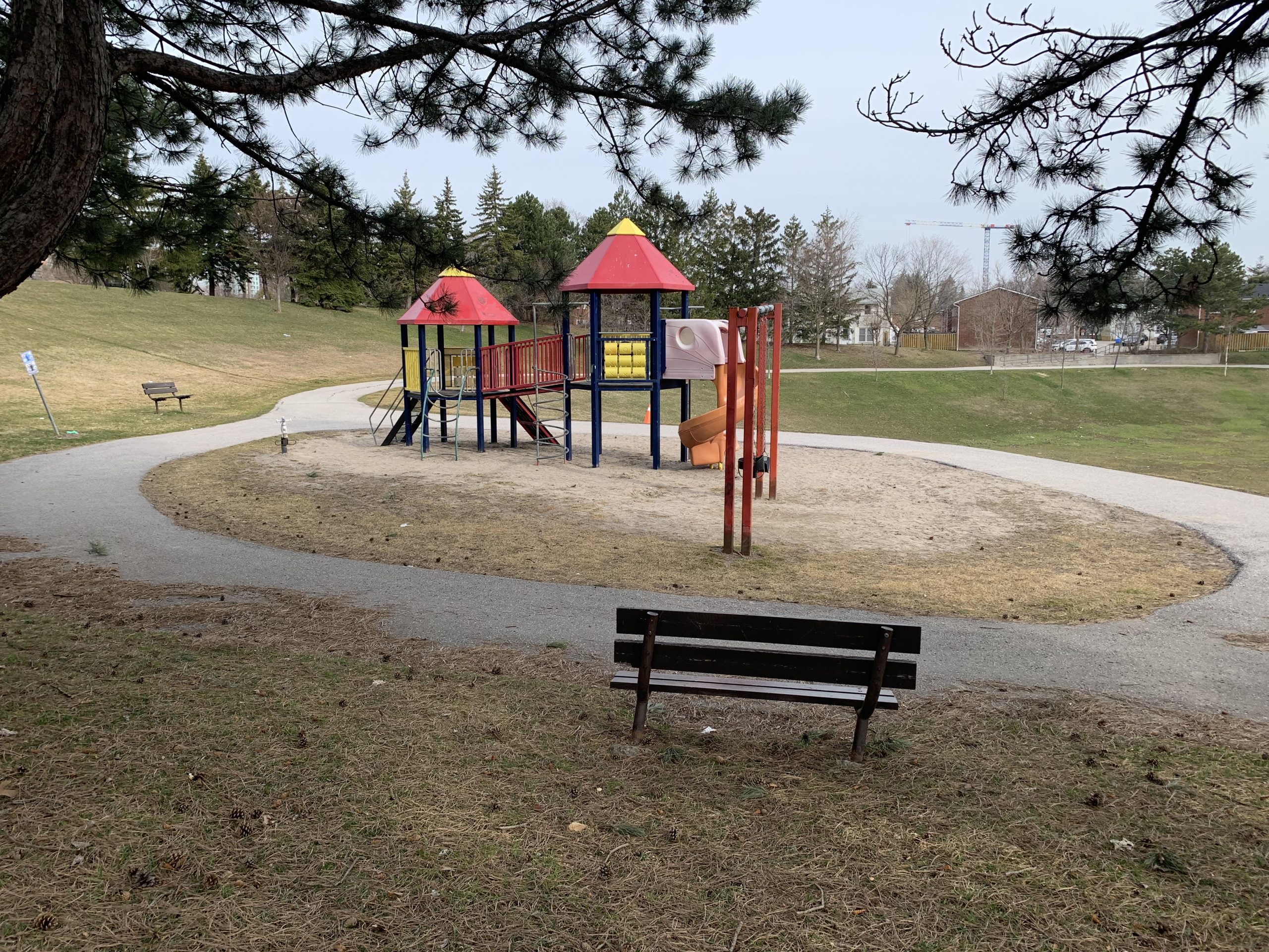 Playground area before improvements looking north. In the foreground is a bench on a grass slope. The playground is surrounded by an asphalt walkway and has sand surfacing. In the background are the grassy slopes of the park with mature trees along the edge.